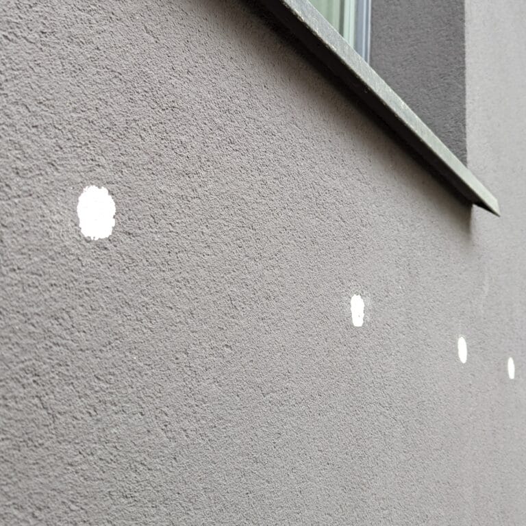 Uszkodzona ciemna elewacja balkonu po siłowym oderwaniu zaczepu siatki wraz z wierzchnią warstwą tynku.