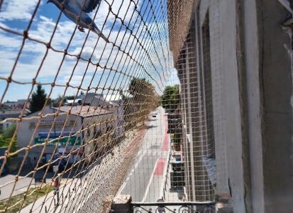Widok na siatkę na balkon przeciw gołębiom, a za nią lecący gołąb, który nie może wlecieć na osiatkowany balkon.